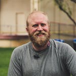 A cheerful bearded man smiling outdoors in a casual setting with greenery.
