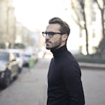 A fashionable man in a black top and glasses poses in an urban street with parked cars.